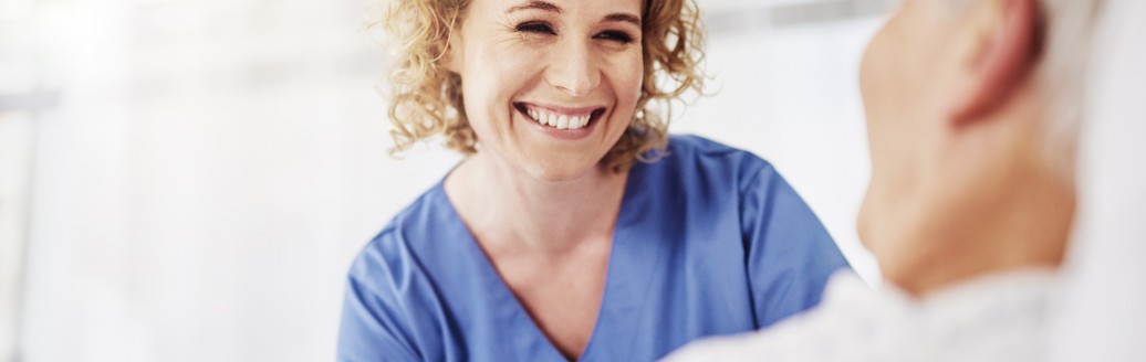 Eine Pflegerin schaut lächelnd einen im Bett liegenden Patienten an. 