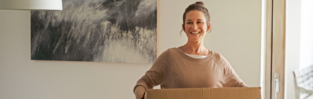 Eine Frau trägt lächelnd einen Umzugskarton durch ein Wohnzimmer.