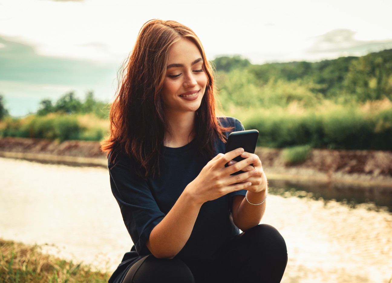 Eine junge Frau schaut am Ufer eines kleinen Flusses auf ihr Smartphone.