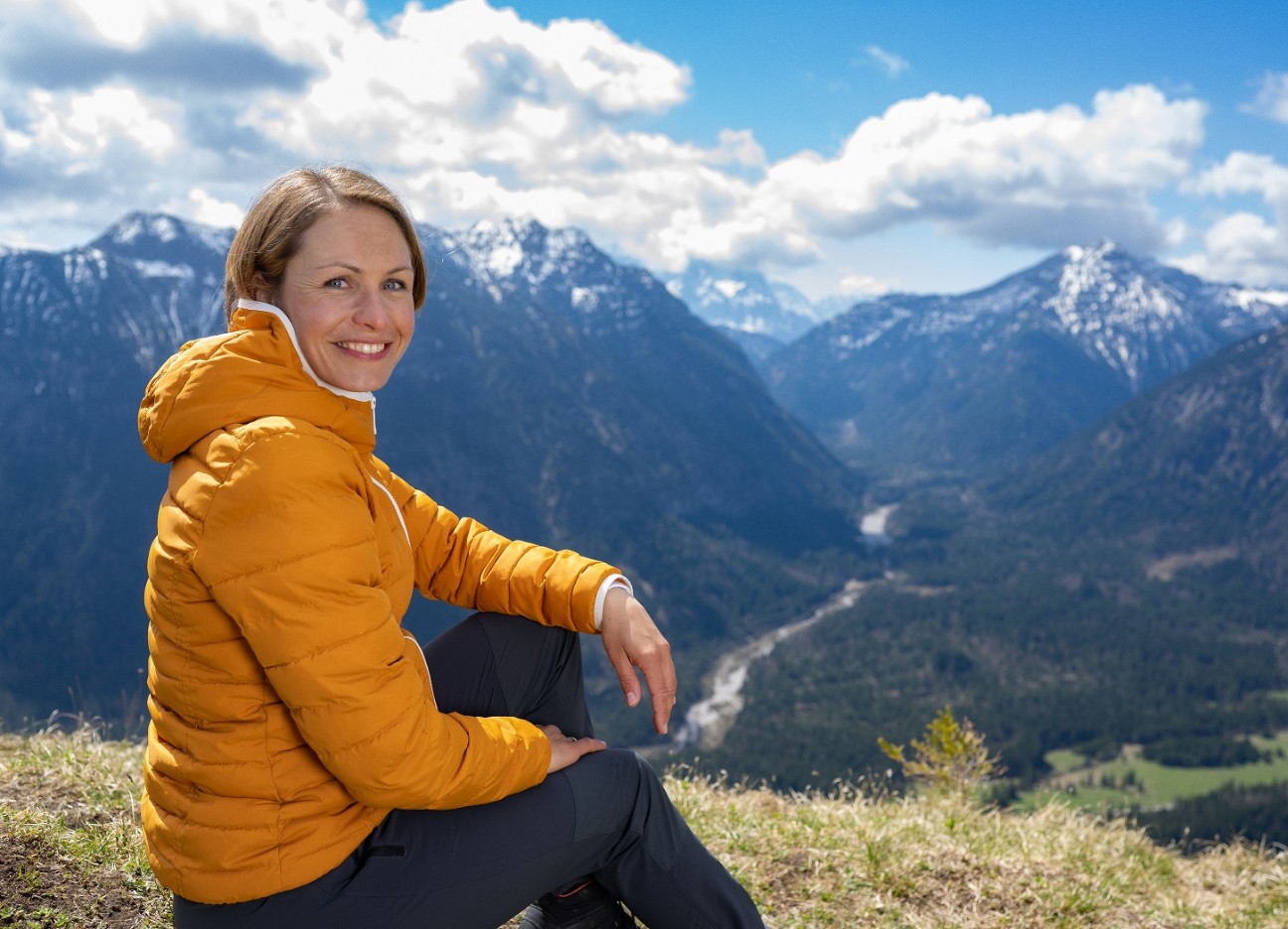 Magdalena Neuner sitzt auf einer Wiese in den Bergen und blickt in die Kamera.