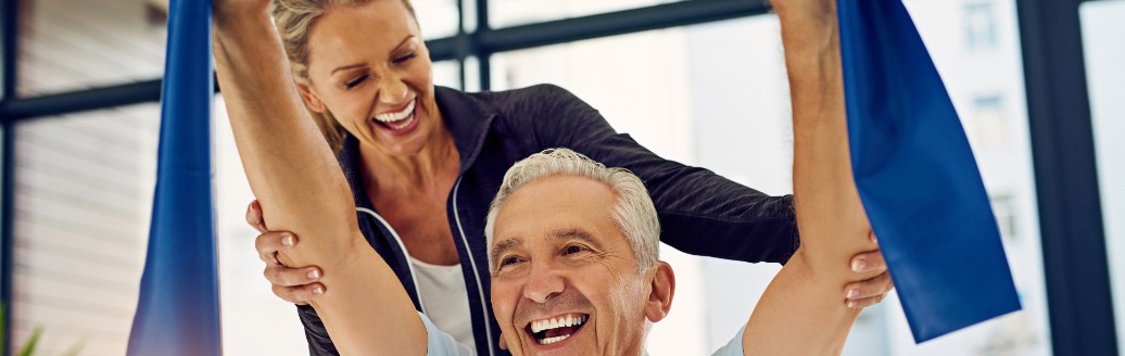Ein glücklicher, älterer Herr führt mit einem Band eine Physioübung aus, während seine Physiotherapeutin ihn im Hintergrund stützt. 