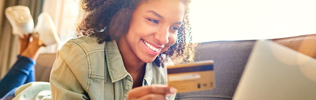 Eine junge Frau liegt auf dem Sofa und tippt lächelnd die Daten ihrer Kreditkarte in ihren Laptop ein.
