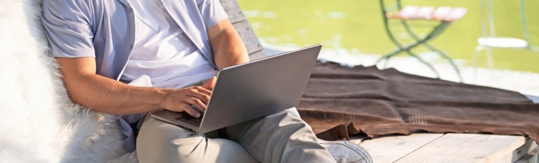 Junger Mann mit T-Shirt und Sommerhemd sitzt an einem See und schaut in ein Laptop