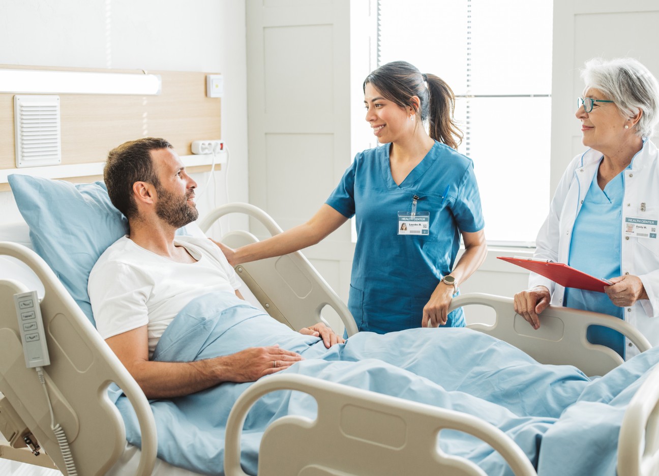 Eine junge Pflegerin und eine ältere Ärztin stehen gemeinsam an einem Patientenbett und unterhalten sich mit dem dort liegenden männlichen Patienten.