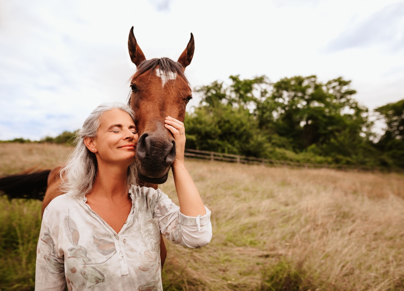 Frau mit heller Bluse lehnt sich an den Kopf eines braunen Pferdes