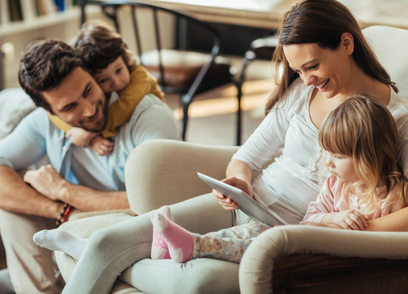 Eine junge Familie, bestehend aus 4 Personen, sitzt zusammen auf dem Sofa. Mutter und Tochter schauen in ein Tablet, Vater und Sohn schauen lächelnd zu ihnen rüber.