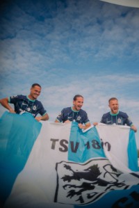 Drei Spieler des TSV 1860 stehen hinter einer Fahne, die sie hochhalten: Zwarts, Sulejmani und Rieder