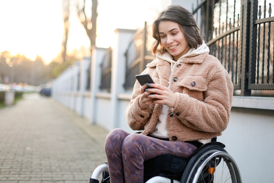 Eine junge Frau, sitzend in einem Rollstuhl, schaut lächelnd auf ihr Smartphone.