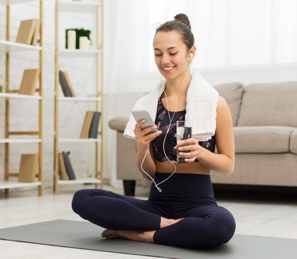 Eine Frau sitzt im Schneidersitz auf einer Sportmatte und lacht in ihr Handy.