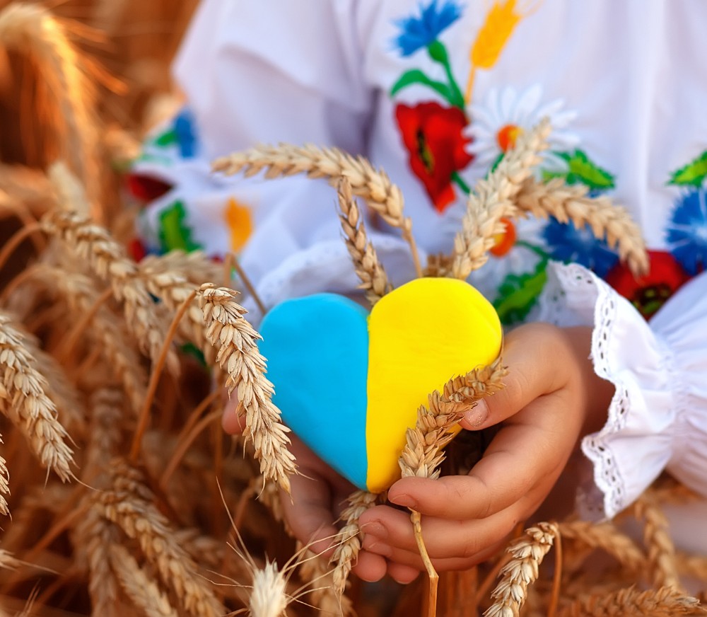 Eine Person in traditioneller Kleidung hält ein blau-gelbes Herz aus Ton inmitten von Weizenähren.