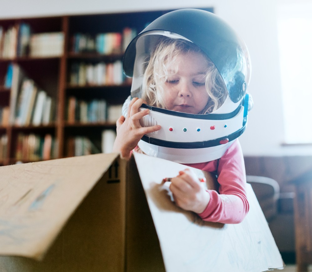 Ein kleines Mädchen sitzt im Wohnzimmer  in einem Umzugskarton und trägt einen Astronautenhelm. Sie malt den Umzugskarton an. 