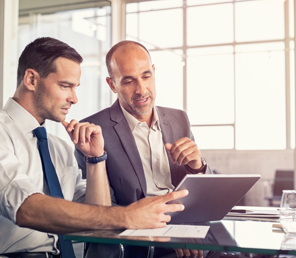 Zwei Business-Männer sitzen zusammen und besprechen etwas am Tablet. 