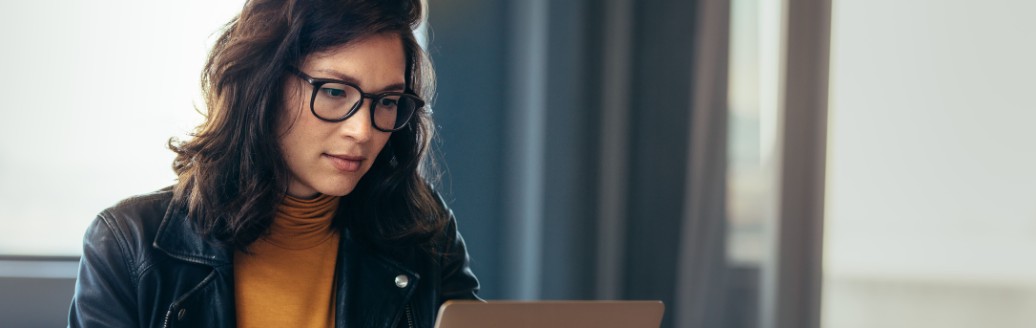 Eine Frau sitzt an einem Tisch und schaut auf ihren Laptop. Neben ihr liegen Dokumente. 