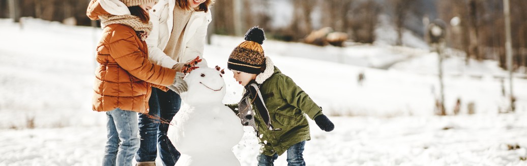 Eine Mutter baut mit ihren zwei Kindern einen Schneemann in einer Winterlandschaft. 
