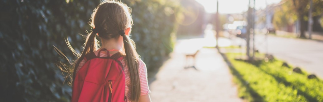 Ein junges Mädchen, fotografiert von hinten, läuft mit ihrem Rucksack eine Straße entlang.