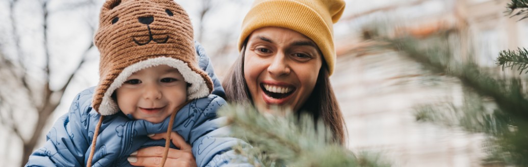 Glückliche Frau hält ihr Baby vor einem Tannenbaum.