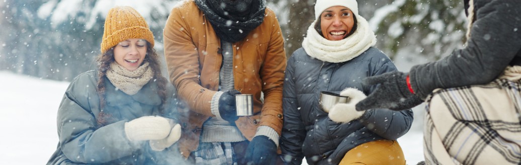 Eine Gruppe von Freunden sitzt in Winterklamotten im Schnee und grillt. 