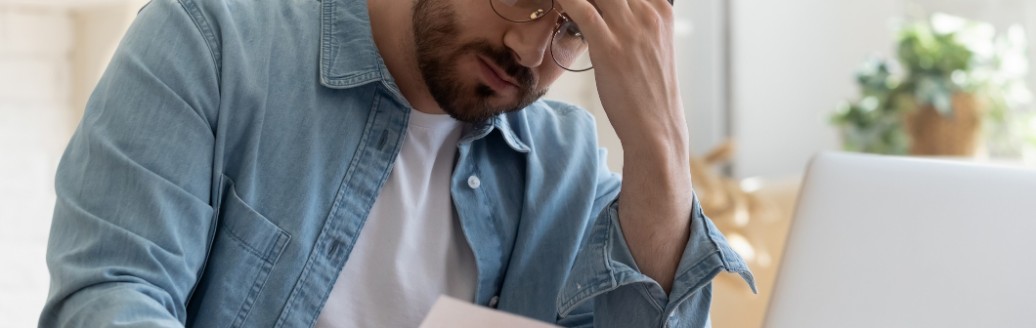 Ein Mann sitzt am Laptop und schaut frustriert auf ein kleineres Blatt Papier. 
