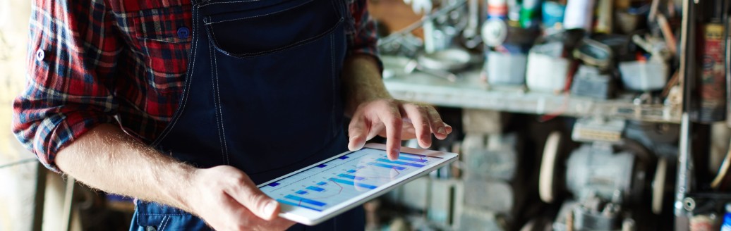 Ein Handwerker steht in einer Garage mit Handwerksequipment und schaut auf ein Tablet. 