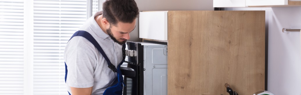 Ein Elektriker steht in einer Küche und untersucht einen Ofen, den er zuvor leicht aus dem Küchenschrank gezogen hat.