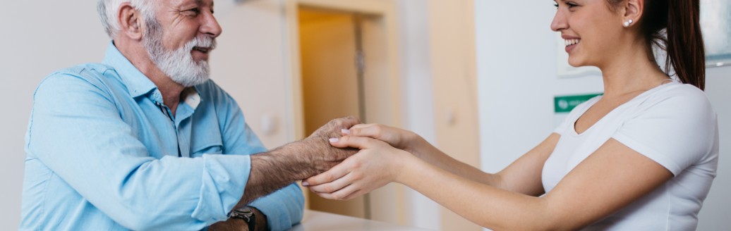 Eine junge Arzthelferin steht hinter der Empfangsrezeption und hält die Hand eines älteren Patienten, der vor ihr am Tresen steht. 