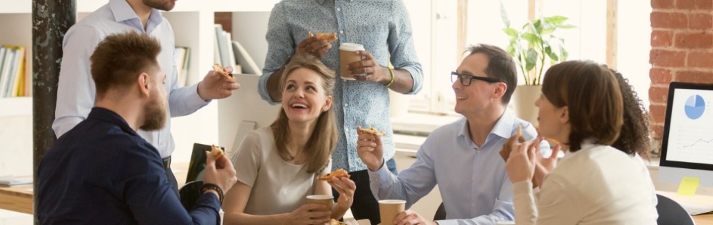 Eine Gruppe von Kollegen sitzt lachend zusammen im Büro an einem Tisch und isst Pizza. 