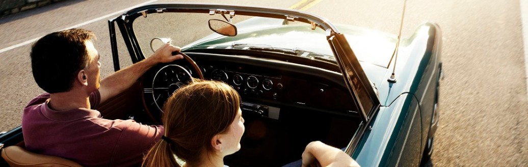 Eine Frau und ein Mann fahren zusammen in einem Oldtimer über eine Landstraße.