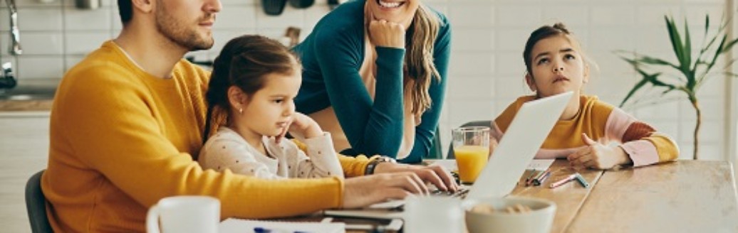 Eine junge Familie sitzt zusammen am Küchentisch. Der Vater arbeitet an seinem Laptop.