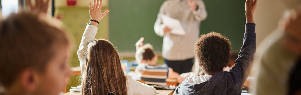 Ein Lehrer steht vor einer Klasse von jungen Kindern an der Tafel und fragt etwas ab. Mehrere Kinder melden sich. 