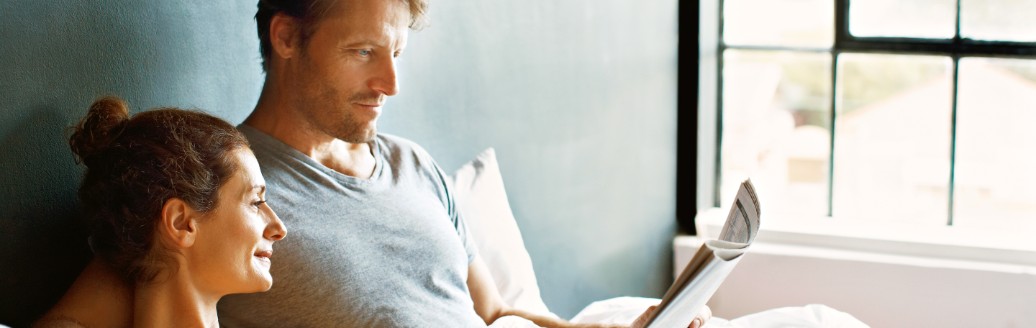Ein Mann und eine Frau liegen zusammen im Bett und lesen sich eine Zeitung durch. Die Frau hält eine Tasse Kaffee in der Hand. 