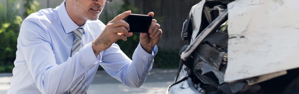 Ein älterer Mann fotografiert mit seinem Mobiltelefon sein Auto, dass einen Schaden erlitten hat.
