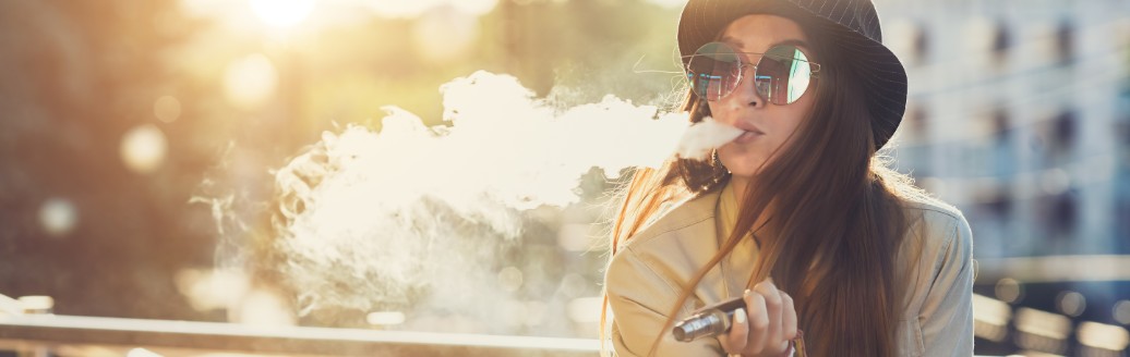 Eine jüngere Frau trägt Hut und Sonnenbrille und raucht eine E-Zigarette.