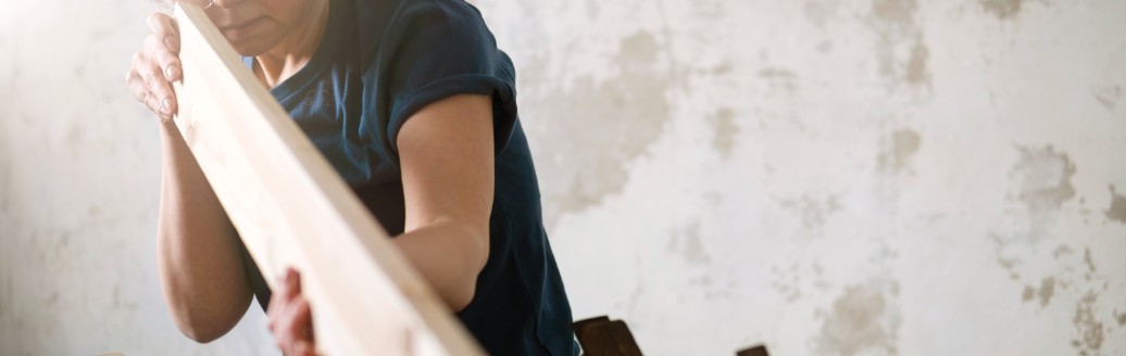 Eine Frau arbeitet an einem länglichen Stück Holz. Sie trägt eine Schutzbrille und neben ihr liegen diverse Werkzeuge. 
