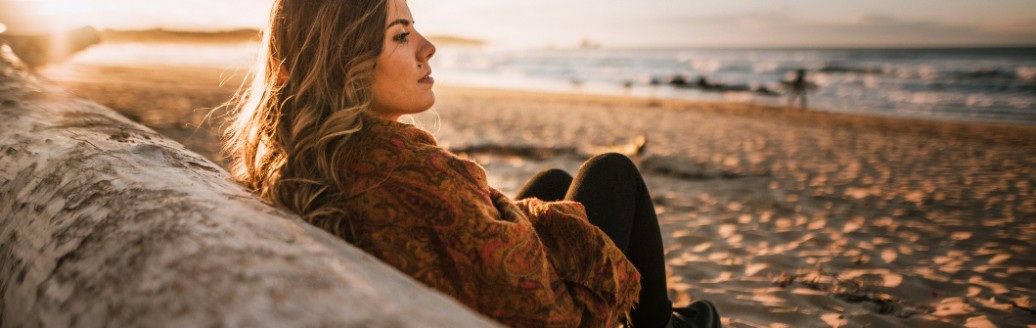 Eine junge Frau sitzt lehnend an einem Holzbalken am Strand. 