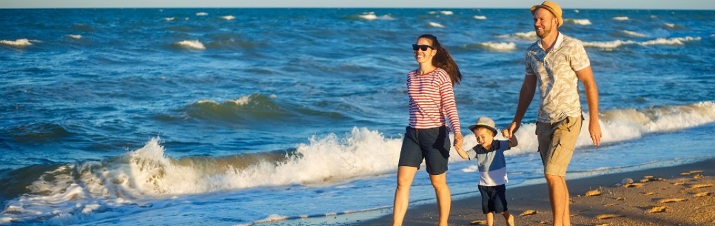 Ein Pärchen läuft mit ihrem Kleinkind Hand in Hand einen Strand entlang. 