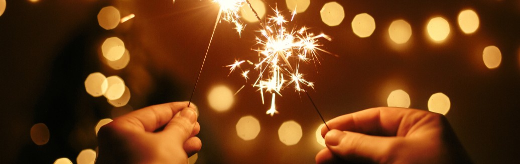 Zwei Hände halten vor einer Lichterkette jeweils eine Wunderkerze.