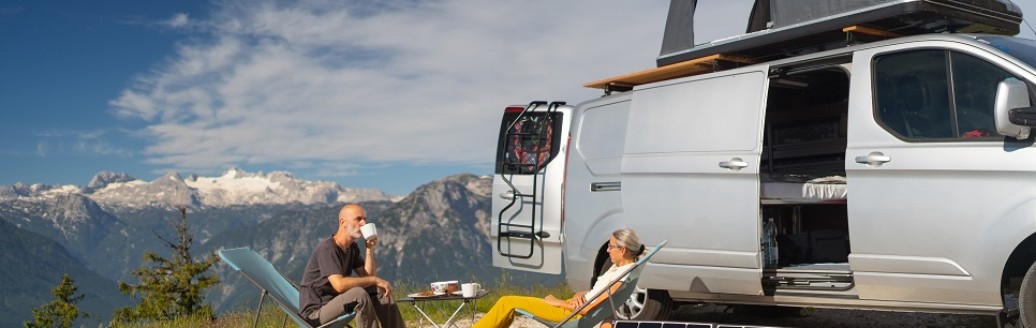 Eine Frau und ein Mann sitzen zusammen in einem Liegestuhl vor ihrem Campervan in den Bergen. Vor ihnen stehen zwei Solarpanels.