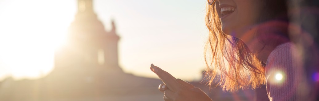 Eine junge Frau schaut lächelnd von ihrem Smartphone hoch, während sie vor einem größeren, weißen Gebäude sitzt. 