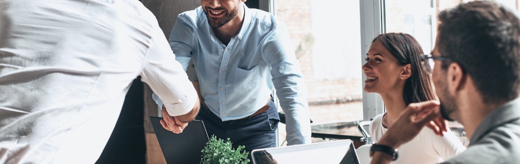 Eine kleinere Gruppe von Leuten sitzen zusammen im Büro und zwei Personen reichen sich die Hände. Sie tragen alle Business-Kleidung. 