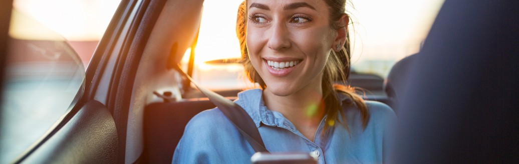 Frau sitzt auf der Rückbank, schaut lachend aus dem Fenster hinaus während sie ein Telefon in der Hand hält.