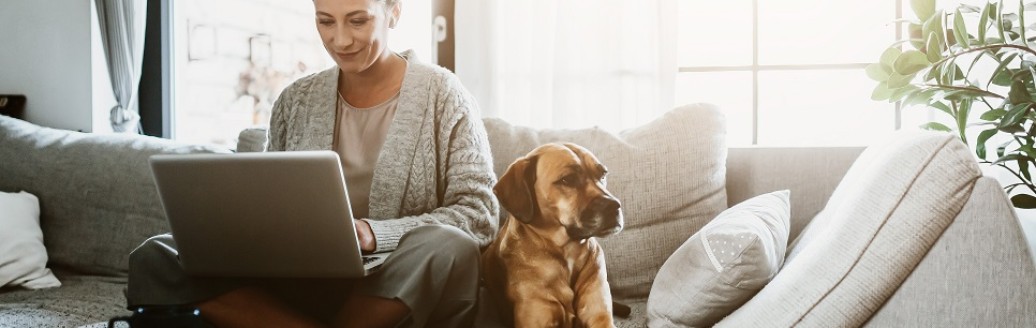 Eine Frau sitzt mit ihrem Laptop auf dem Sofa. Neben ihr liegt ihr Hund. 