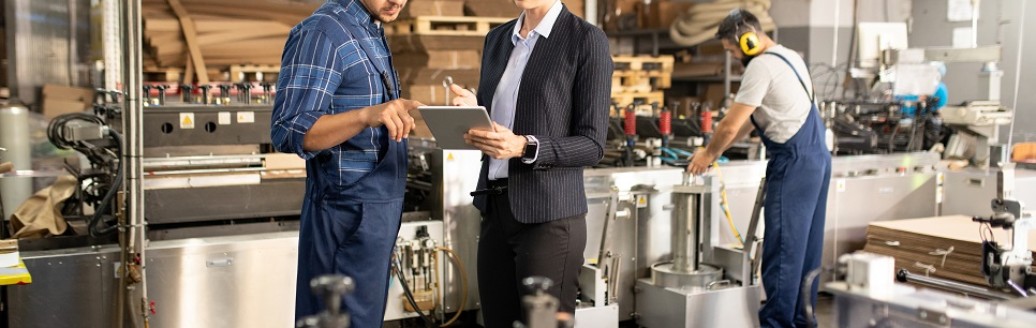Eine Business-Frau und ein Bauarbeiter sprechen zusammen über ein Dokument, welches die Frau in der Hand hält. Die Umgebung ist eine Fabrik. 