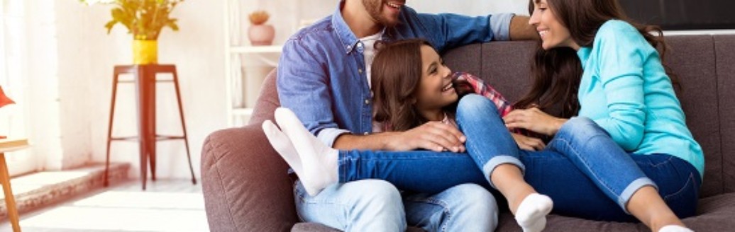 Zwei junge Eltern sitzen lachend mit ihrer Tochter auf dem Sofa.