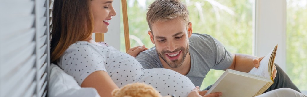 Eine hochschwangere Frau liegt aufrecht im Bett, neben ihr ein Teddybär und ein Mann liest ein Buch vor, während er auf den Babybauch schaut. 