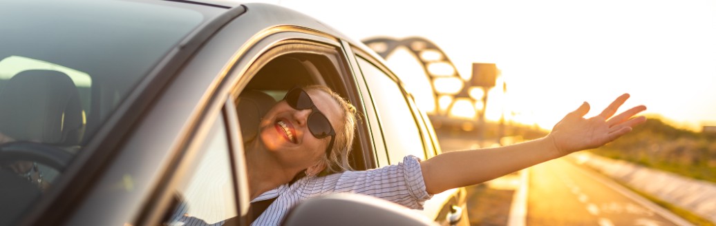 Eine junge blonde Frau fährt in glücklich in der Sonne ihr Auto, trägt eine Sonnenbrille, streckt einen Arm aus dem Fahrerfenster und lacht.
