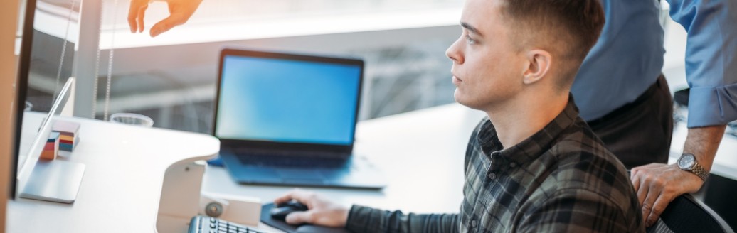 Ein junger Mann sitzt am PC und ein älterer Mann zeigt ihm etwas. 