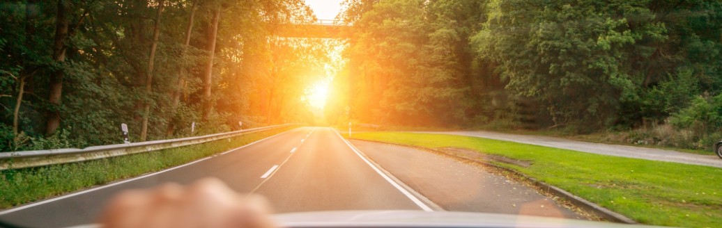 Eine zusehende Hand am Lenkrad eines Autos. Das Auto fährt eine Landstraße im Sonnenuntergang entlang. 