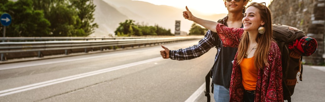 Zwei Backpacker stehen mit ihren Rucksäcken am Straßenrand und halten ihren Daum raus. Sie warten auf eine Mitfahrgelegenheit. 