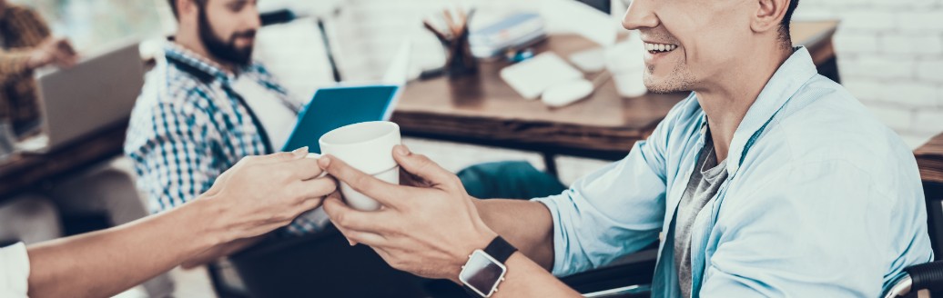 Ein junger Mann sitzt im Rollstuhl und ihm wird eine Tasse Kaffee gereicht. Mit ihm sitzt ein Kollege im Büro. 