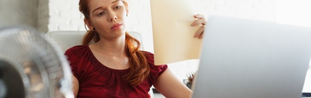 Eine Frau sitzt verschwitzt am Laptop, vor ihr ein Ventilator und sie fächert sich Luft mit Unterlagen zu. 